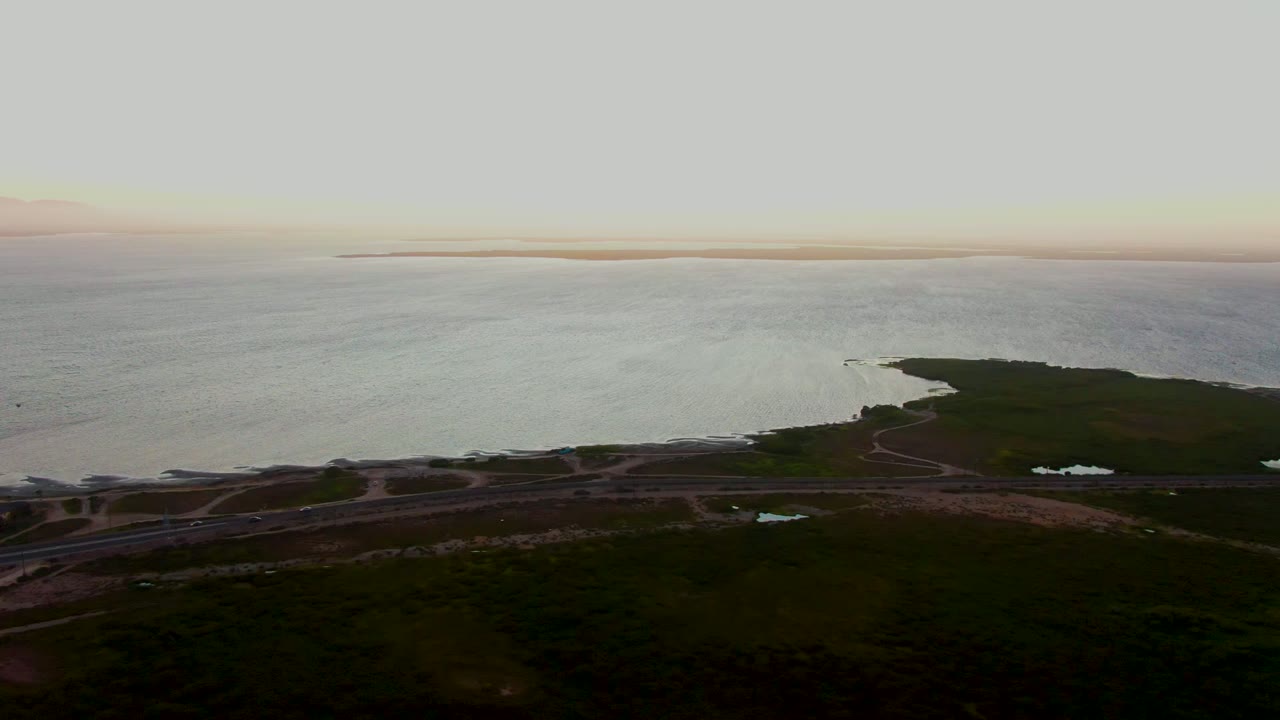 Aerial frame over San Carlos, Baja California Sur shows rippled coastal water beyond dark green shoreline and roads, with a pale horizon and no visible people.