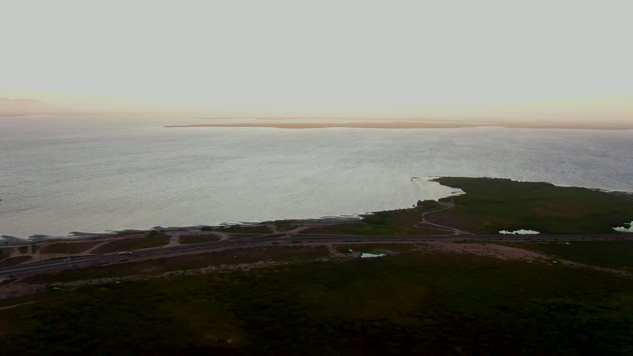 Wide aerial coastal view with water ripples, curved shoreline, roads, and small structures.