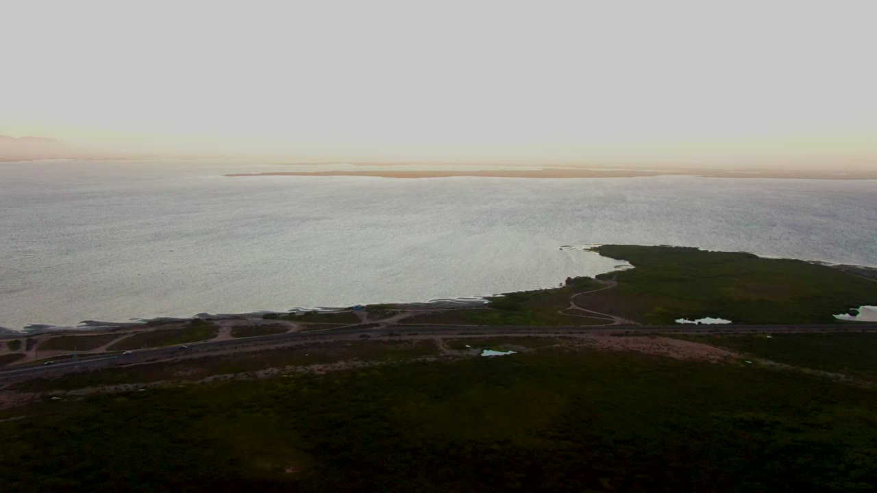 Aerial view over San Carlos, Baja California Sur shows dark foreground vegetation, a road crossing the coast, a green point of land, and calm rippled ocean under soft light, with no visible people.