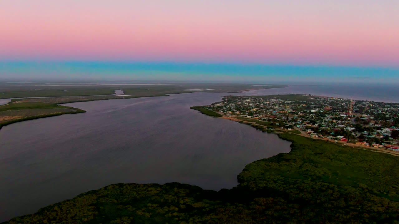 Aerial dusk view over San Carlos, Baja California Sur, with dark forest in the foreground, a wide estuary, shoreline buildings, a flat horizon, and no visible people.