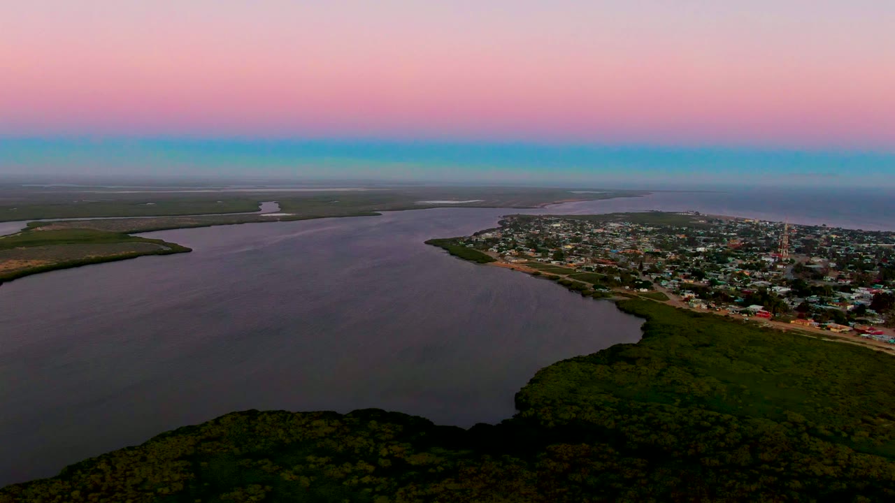 Wide water and shoreline town seen from above with forest below and dusk color across the sky.