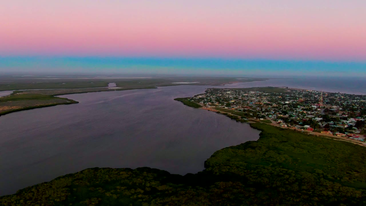 Aerial frame from San Carlos, Baja California Sur, with dark rippled estuary water, compact shoreline buildings, green vegetation in the lower frame, open distant land, and no visible people.