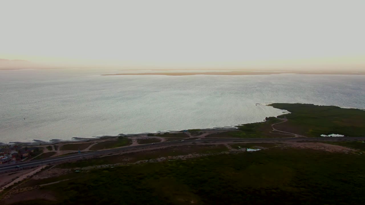 Aerial view over San Carlos, Baja California Sur with a coastal road in the foreground, dark green shoreline reaching into calm ocean, distant horizon, and no visible people.