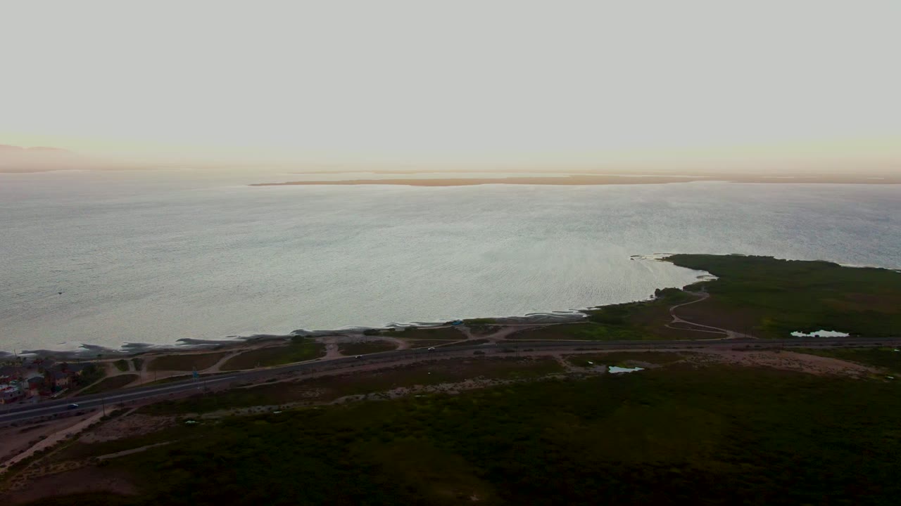 Wide aerial shoreline with ocean, road, vegetation, and distant land on the horizon.