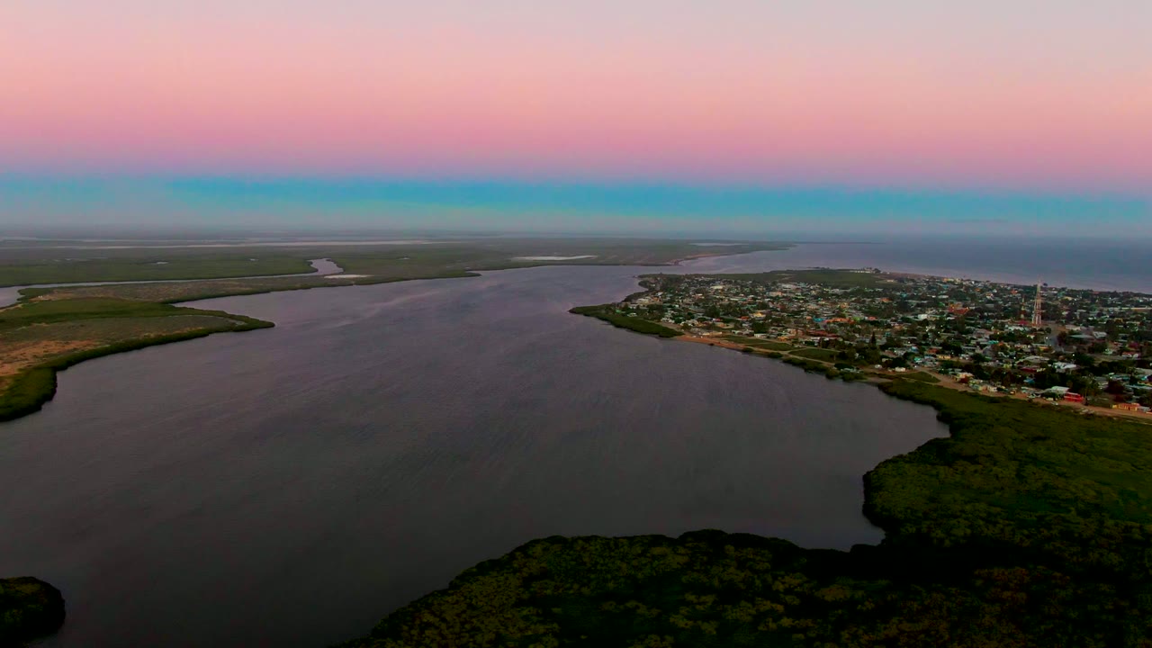 Aerial dusk view over San Carlos, Baja California Sur, with broad coastal water in front, green shoreline at the edges, a small town on the peninsula, pink-blue sky, and no visible people.