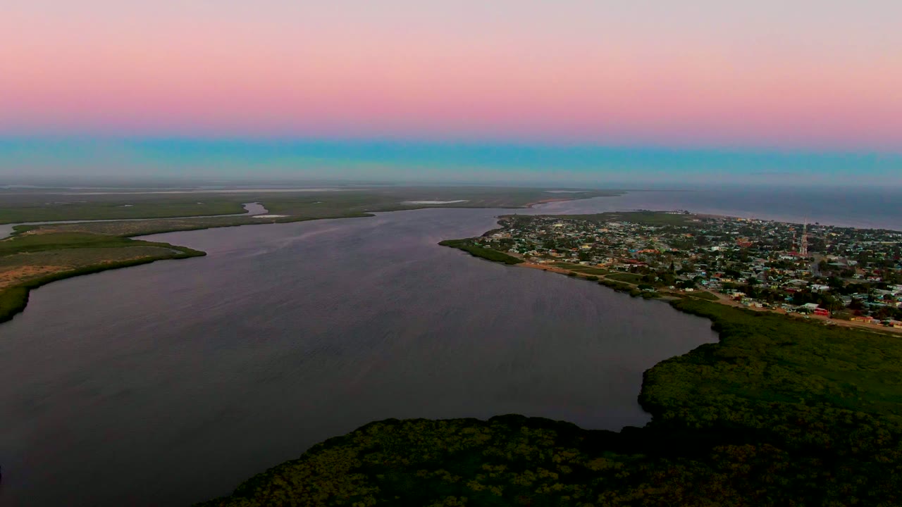 Aerial view of San Carlos, Baja California Sur, with calm coastal water across the foreground, the town clustered to the right, green land to the left and bottom, a blue horizon band below pink sky, and no visible people.