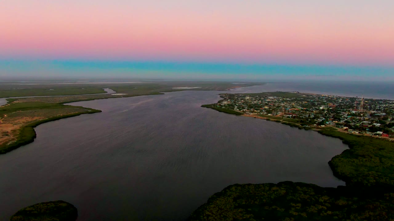 Wide aerial view of water curving around a vegetated shore with a town beyond at dusk.