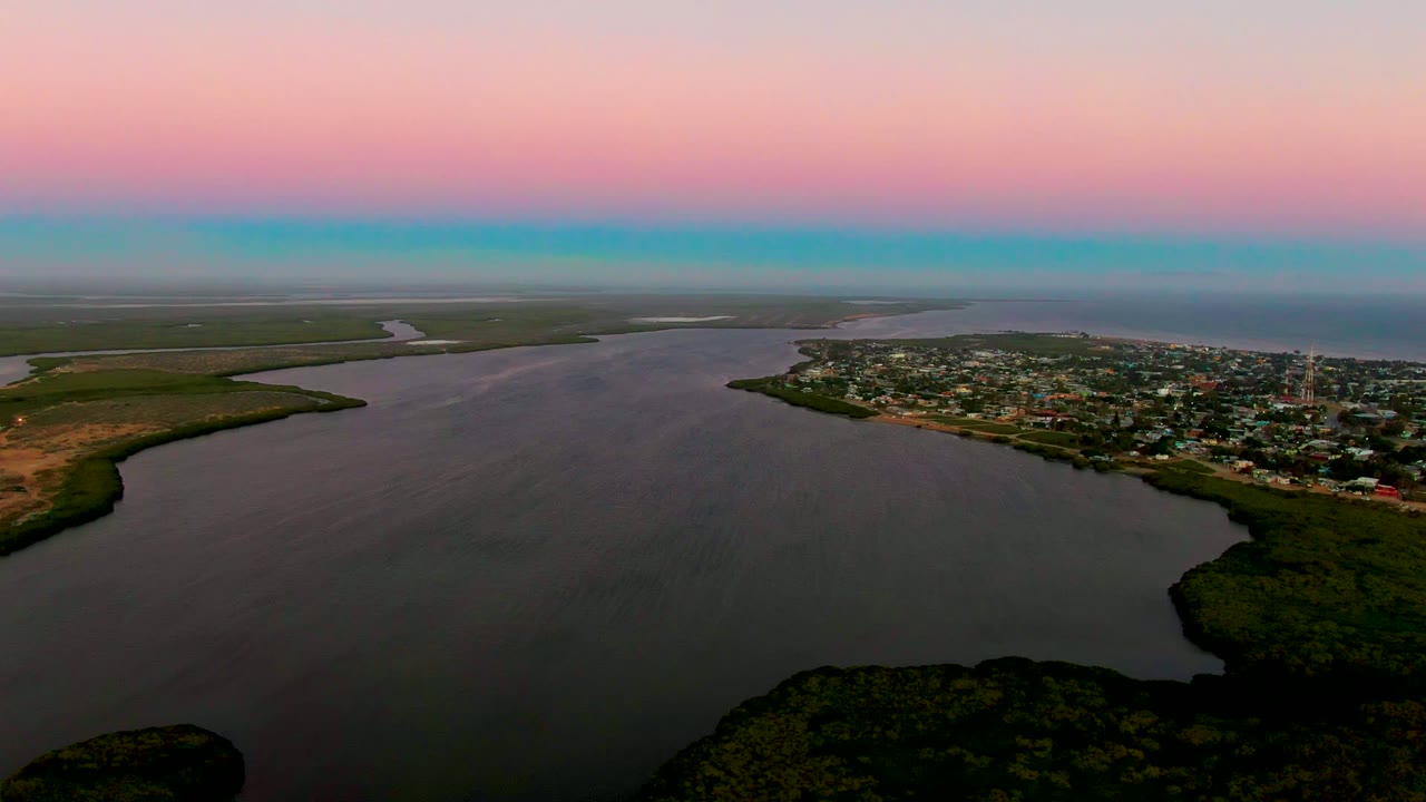 Drone-height view of San Carlos, Baja California Sur with calm water occupying most of the frame, a compact town on the right shoreline, low green banks, and no visible people.