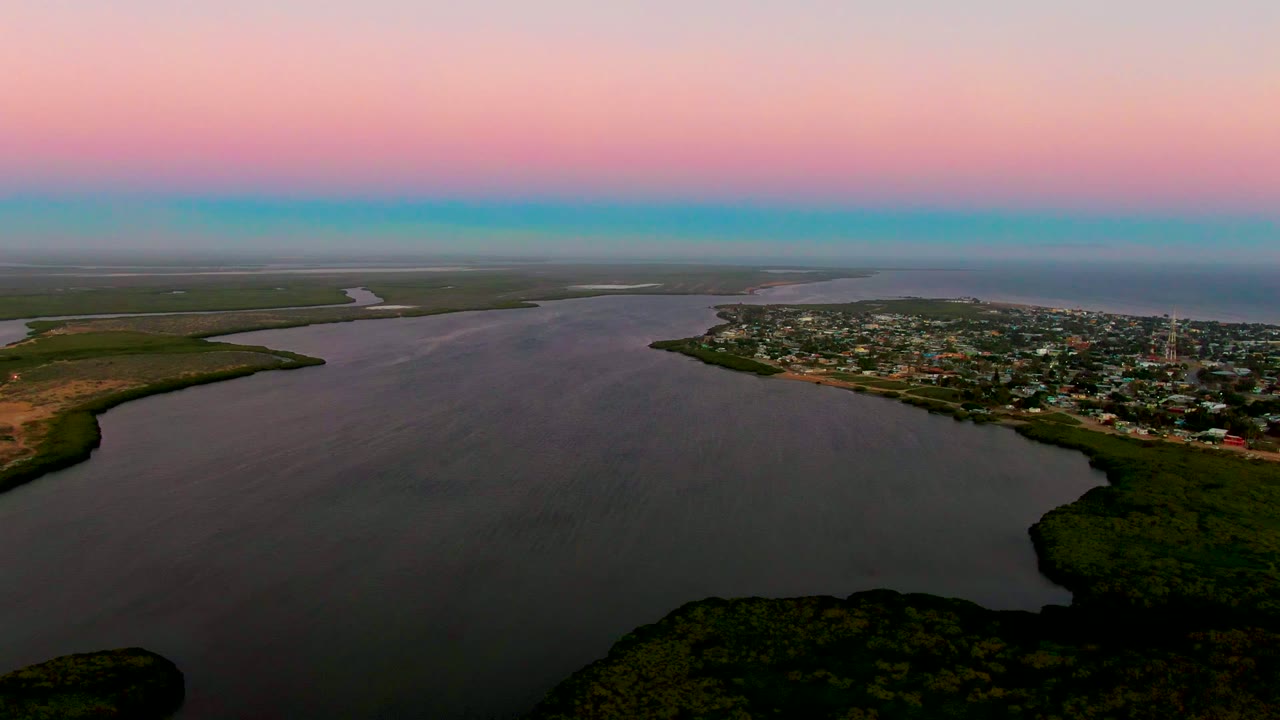 Dusk aerial over a river mouth or inlet with green banks and town buildings on the right.