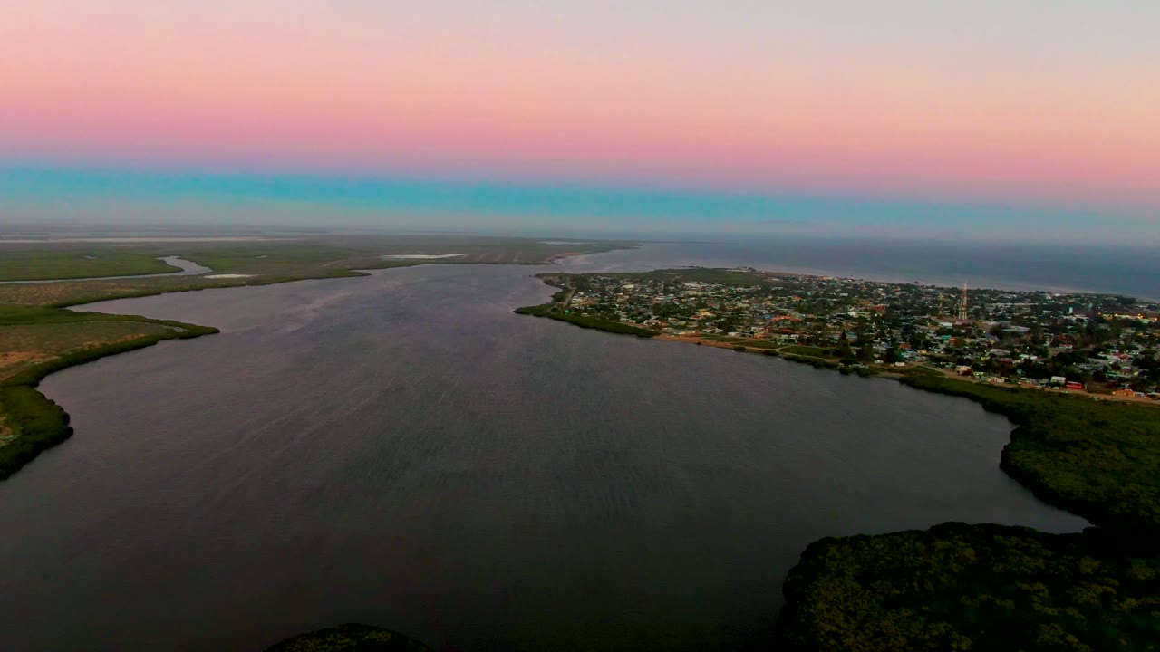 Opening aerial frame over San Carlos, Baja California Sur, with calm estuary water in front, a coastal town to the right, green shoreline to the left, pink-blue sunset sky above, and no visible people.