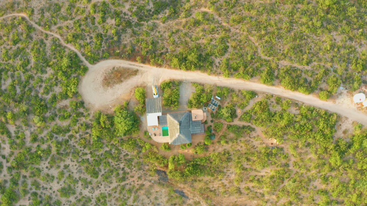 Overhead view in Cabo Pulmo, Baja California Sur of a pale dirt road, a small rural compound, a turquoise rectangular pool or tank, white roadside objects, surrounding brush, and no visible people.
