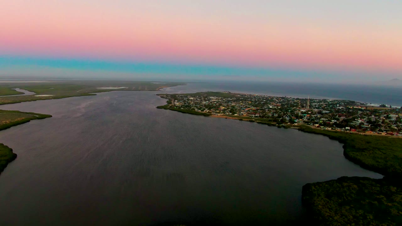 Aerial dusk view over San Carlos, Baja California Sur, with wide dark water in the foreground, green shoreline edges, town buildings along the right shore, pink and blue sky, and no visible people.