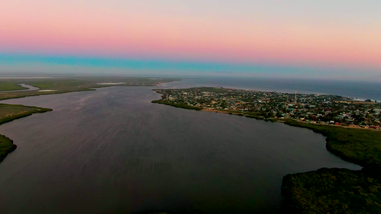 Wide aerial over river water, green shoreline, and town buildings beneath a pink dusk sky.