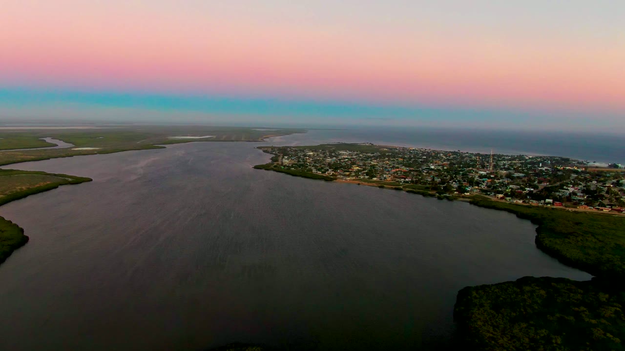 A high aerial view looks across the dark river surface toward San Carlos, with clustered buildings near the shore, greenery at the lower right, a distant coast, and no visible people.