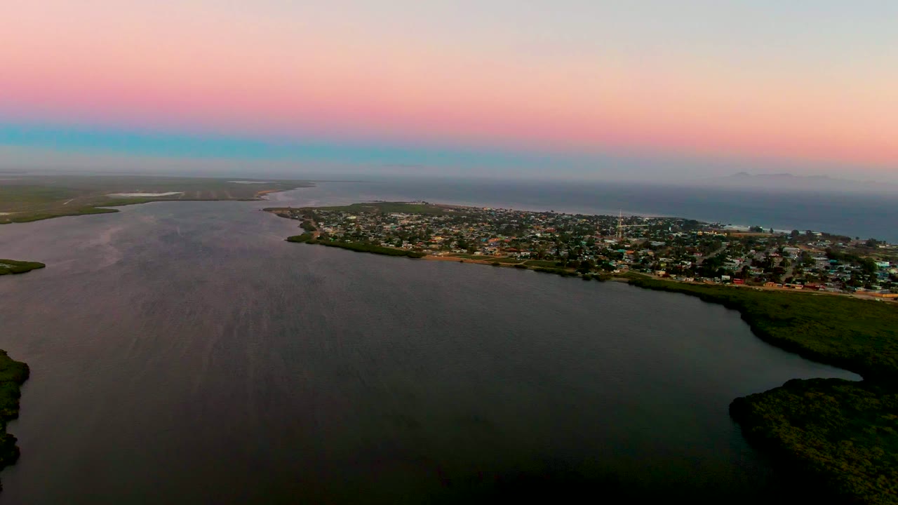 Aerial dusk frame over San Carlos, Baja California Sur, showing calm inlet water in the foreground, a shoreline town across the middle, dark green vegetation, distant ocean, and no visible people.
