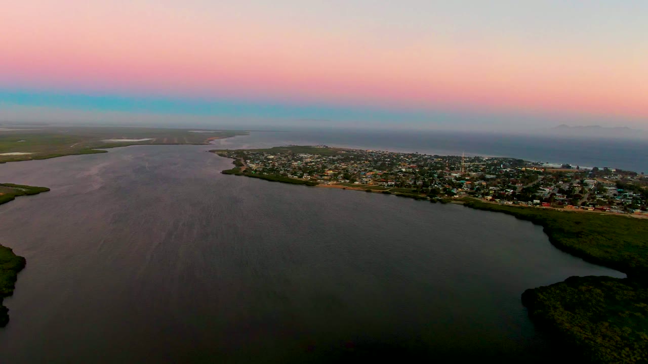 The 0:02 frame shows the San Carlos shoreline from above, with smooth inlet water leading to low buildings, green shore edges, a distant ocean horizon, and no visible people.
