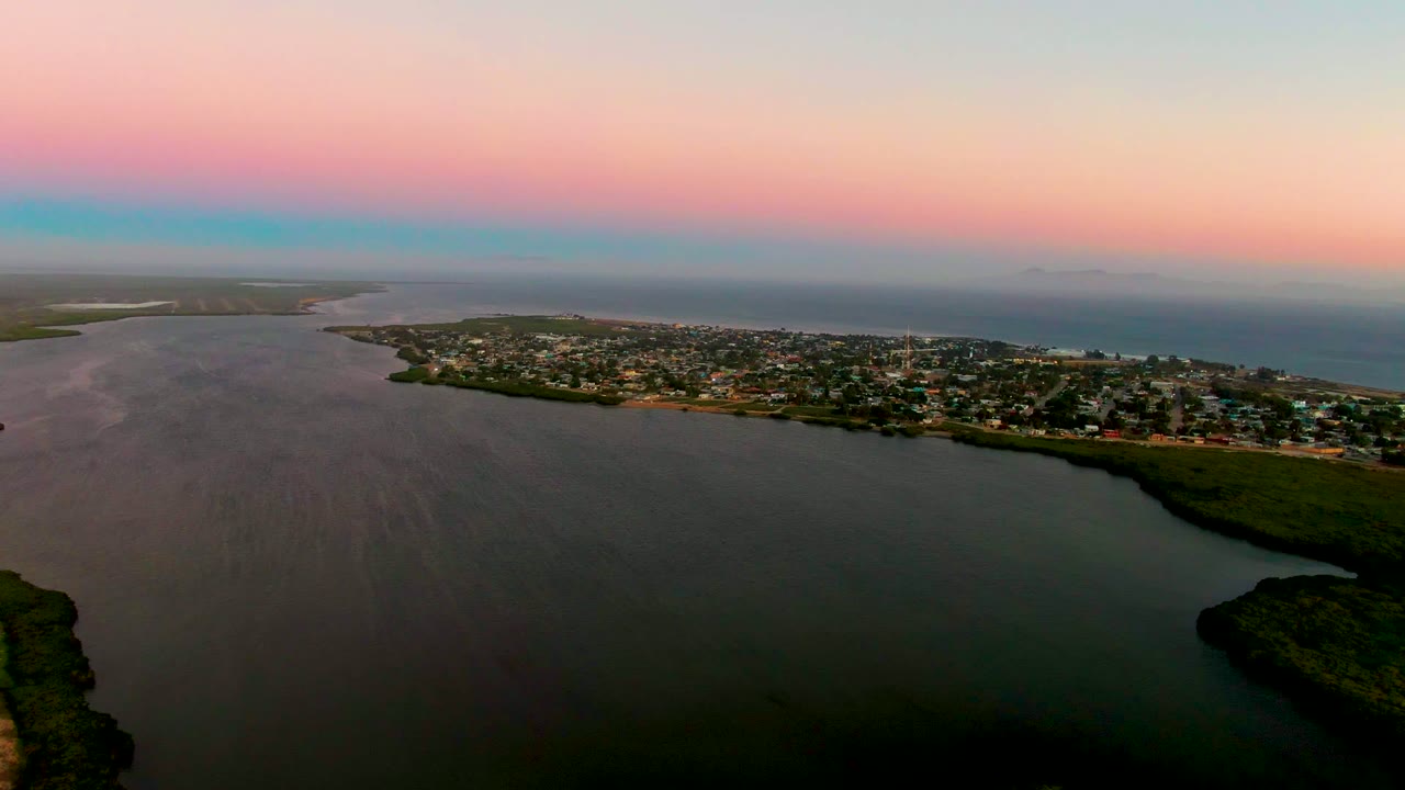 Aerial view over San Carlos, Baja California Sur, showing calm water in the foreground, a shoreline town in the midground, ocean on the horizon, pink and blue sunset sky, and no visible people.
