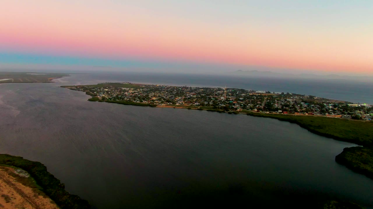 Opening aerial view over San Carlos, Baja California Sur, with dark lagoon water in the foreground, town buildings along the shoreline, ocean beyond, and no visible people.