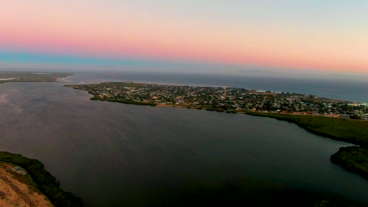 Wide aerial view of coastal buildings between lagoon and ocean under a pastel dusk sky.