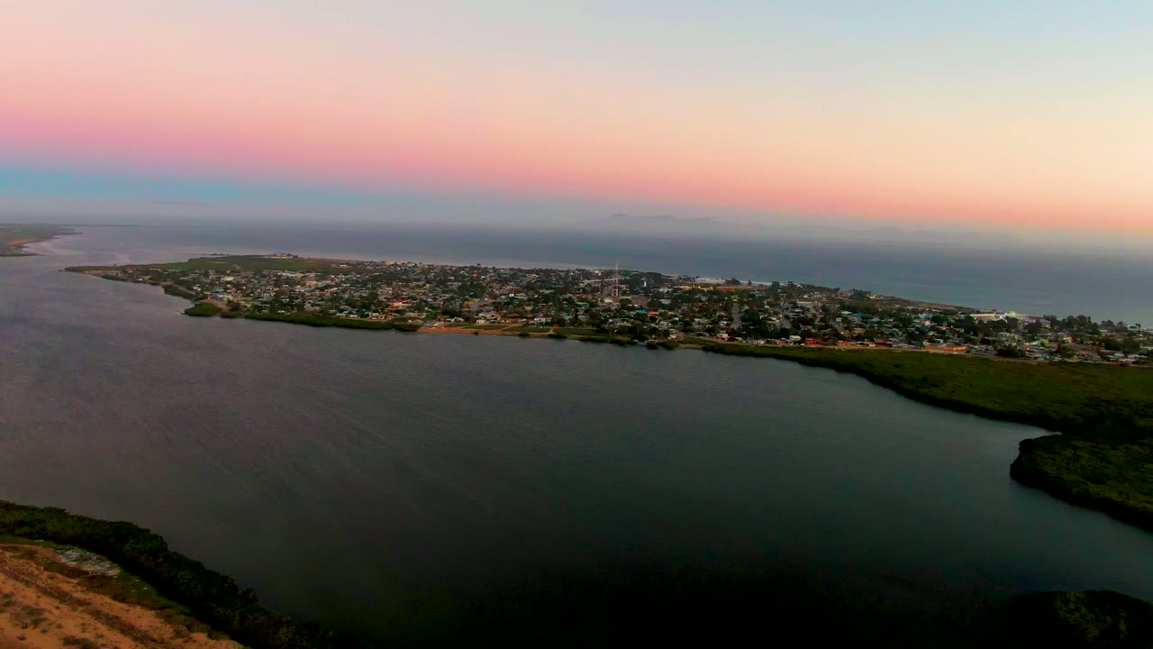 Aerial dusk view over San Carlos, Baja California Sur, with dark lagoon water in the foreground, shoreline greenery, a low town across the middle, ocean beyond, and no visible people.