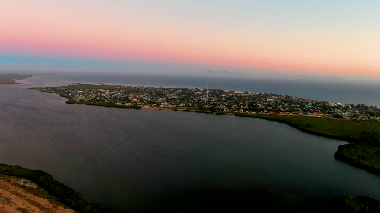 Wide aerial over lagoon water and a coastal town beneath a pastel dusk sky.