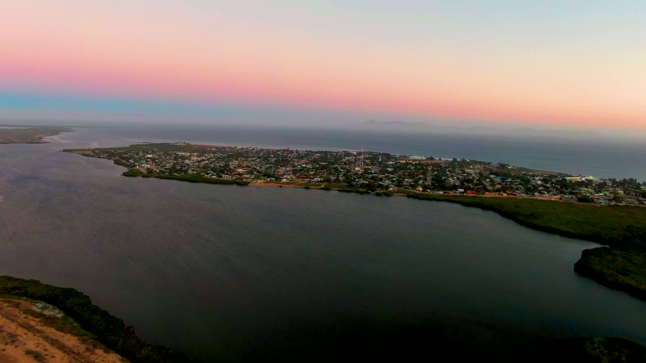 Aerial view of San Carlos, Baja California Sur, with foreground lagoon water, a green shoreline edge, clustered low buildings, ocean in the background, and no visible people.