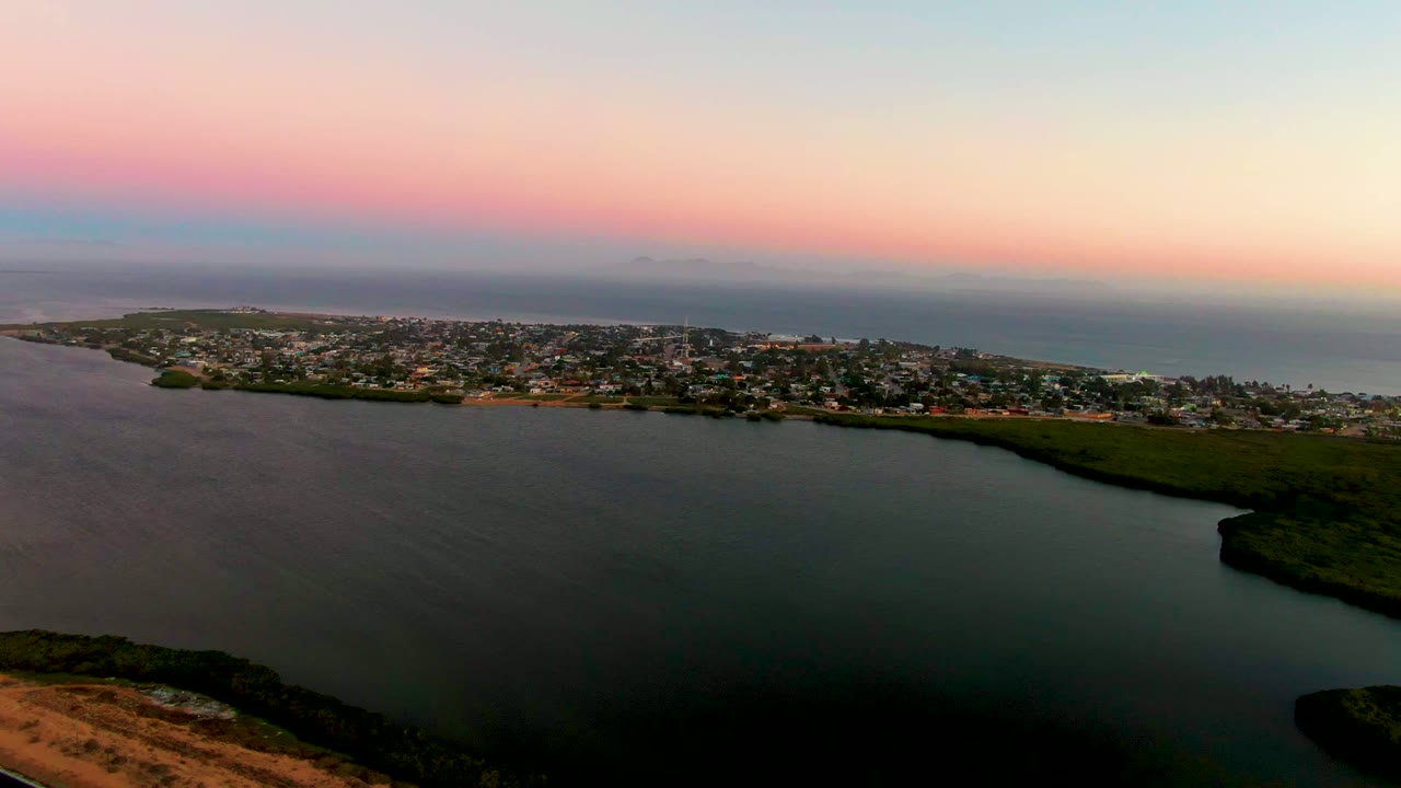 At 0:02, the aerial view shows San Carlos stretched along the shoreline, dark water filling the lower frame, open ocean behind the town, and no visible people.