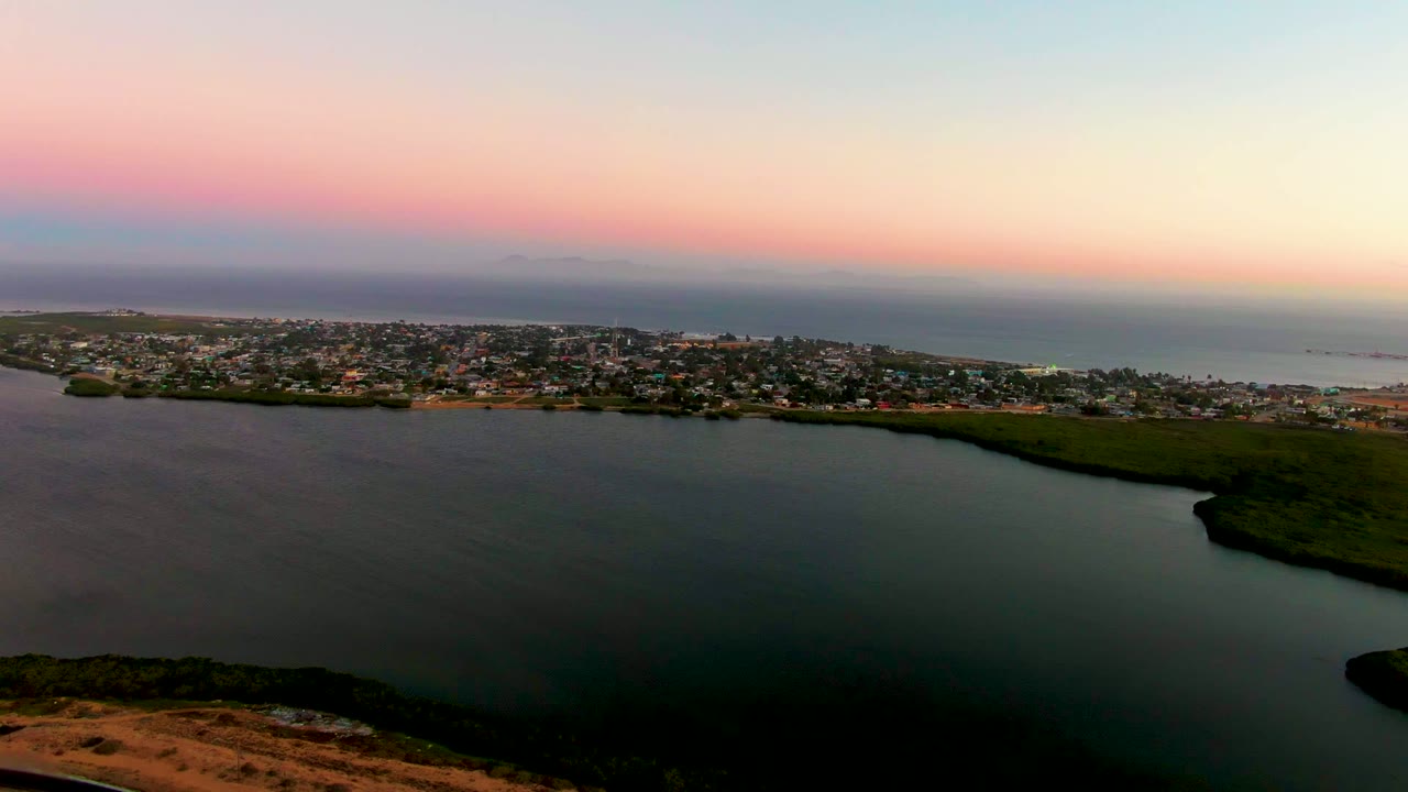 Opening frame of San Carlos, Baja California Sur from above, with dark lagoon water in front, a coastal town across the shore, the ocean beyond, and no visible people.