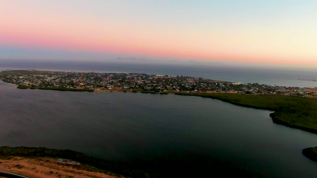 Wide aerial view over lagoon water toward a low town and ocean at dusk.
