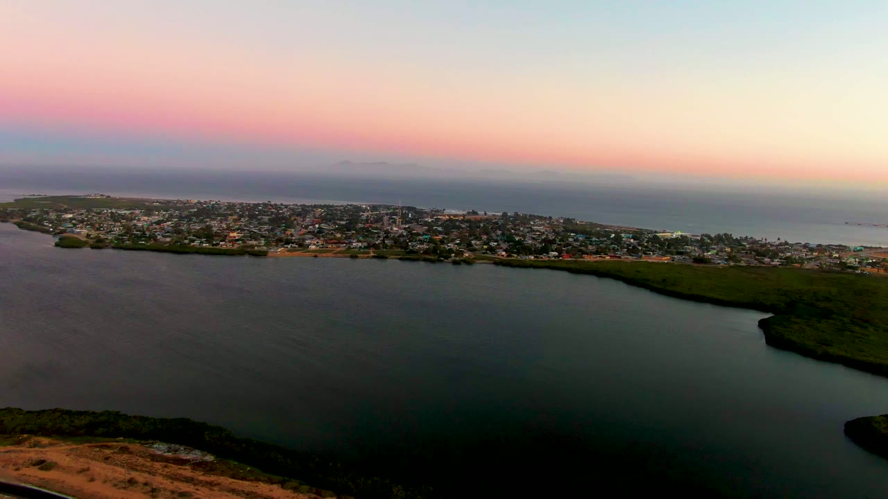 Mid-early frame of San Carlos, Baja California Sur with the lagoon occupying the lower frame, clustered buildings along the shore, ocean behind them, and no visible people.