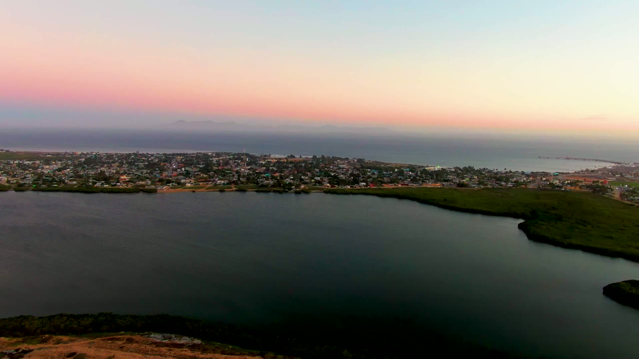 Aerial view over San Carlos, Baja California Sur with a dark lagoon in the foreground, coastal buildings on the far shore, ocean beyond, pink-blue sunset sky, and no visible people.