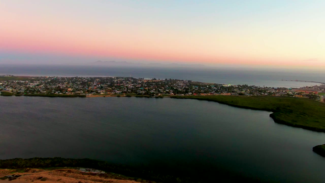 A wide aerial frame shows San Carlos, Baja California Sur along the far edge of a calm lagoon, with darker green land at right, ocean beyond, pastel sunset color, and no visible people.