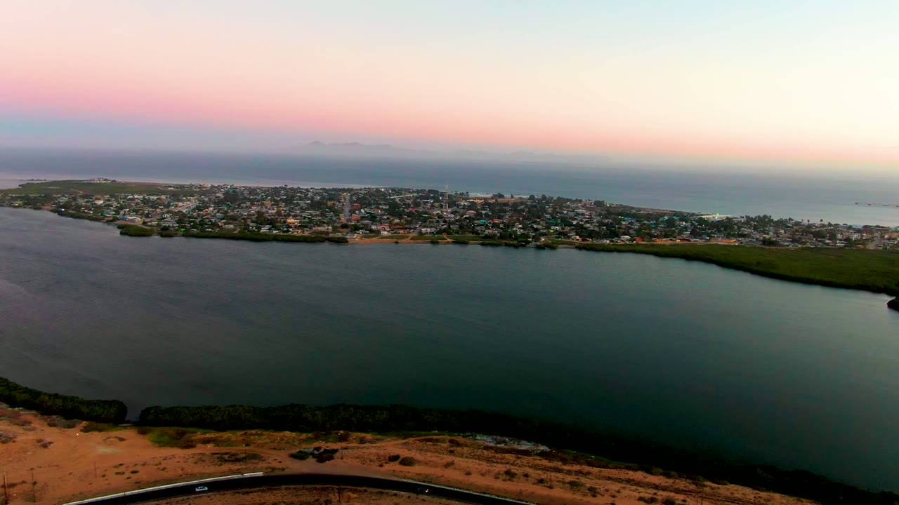 Opening aerial frame over San Carlos, Baja California Sur, with calm foreground water, a shoreline road at the bottom edge, the coastal town across the middle, pink-blue sky, and no visible people.