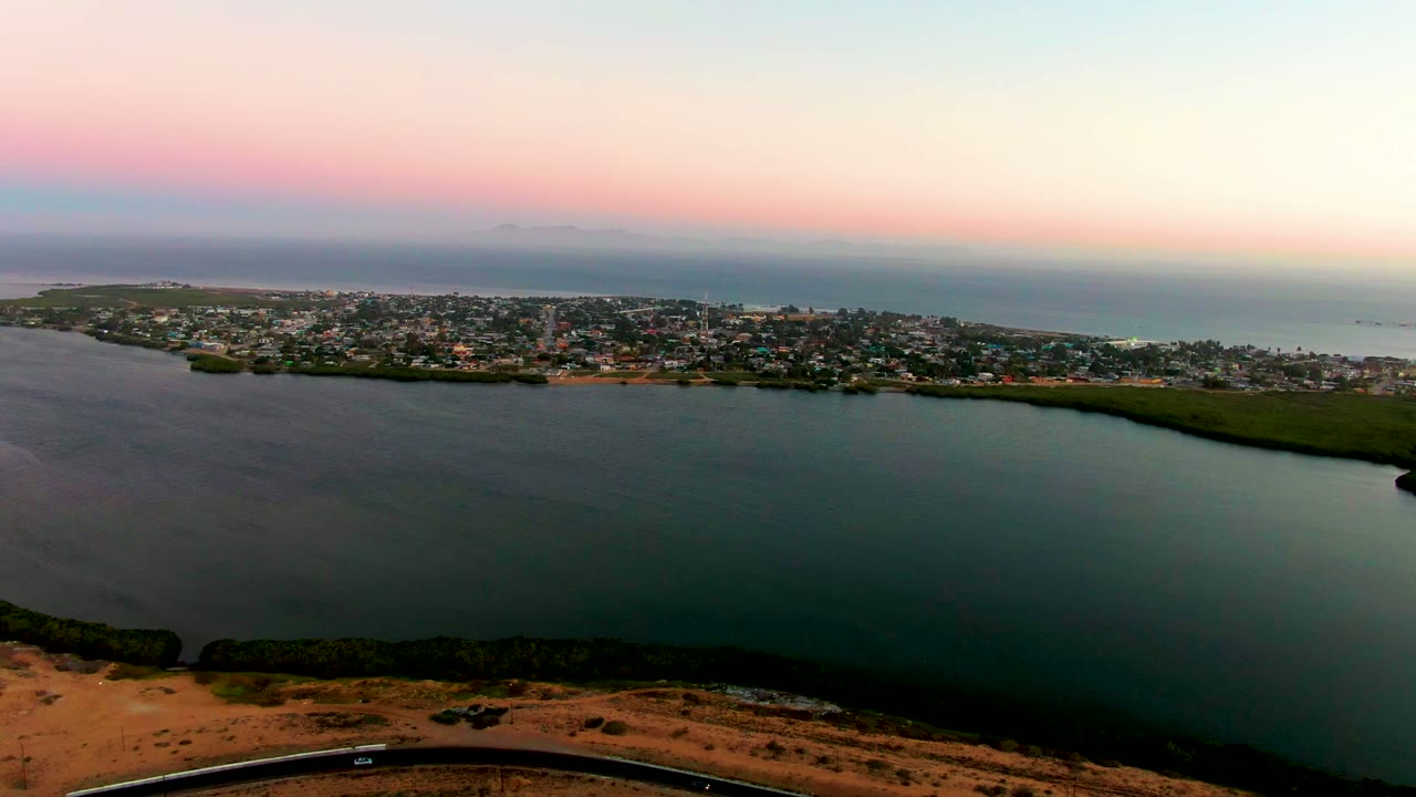 Aerial frame over San Carlos, Baja California Sur, with the shoreline road slightly more centered along the bottom, dark water in the foreground, town buildings in the midground, and no visible people.