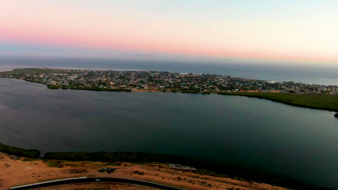 Aerial view of San Carlos, Baja California Sur, showing broad foreground water, the town running across the horizon line, a thin shoreline road at the bottom, soft sunset color, and no visible people.
