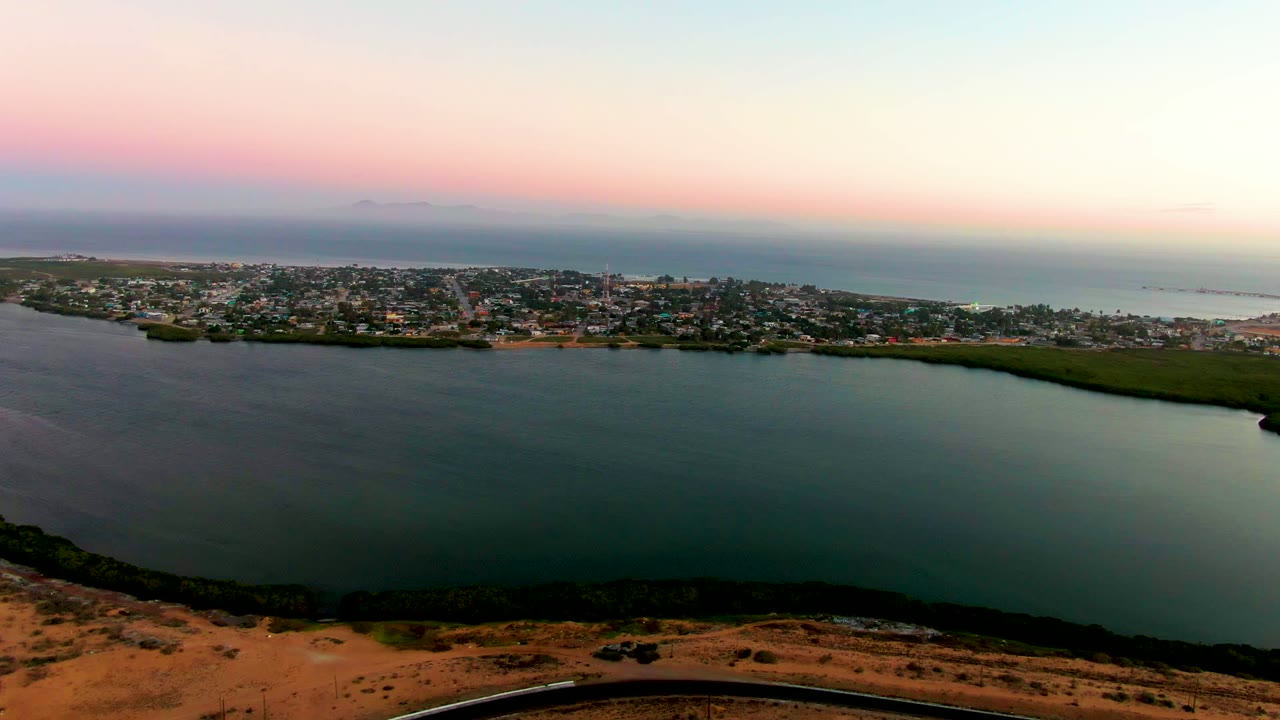 Aerial view over San Carlos, Baja California Sur, with calm bay water in the foreground, town buildings across the shore, ocean beyond, pink sunset sky, and no visible people.