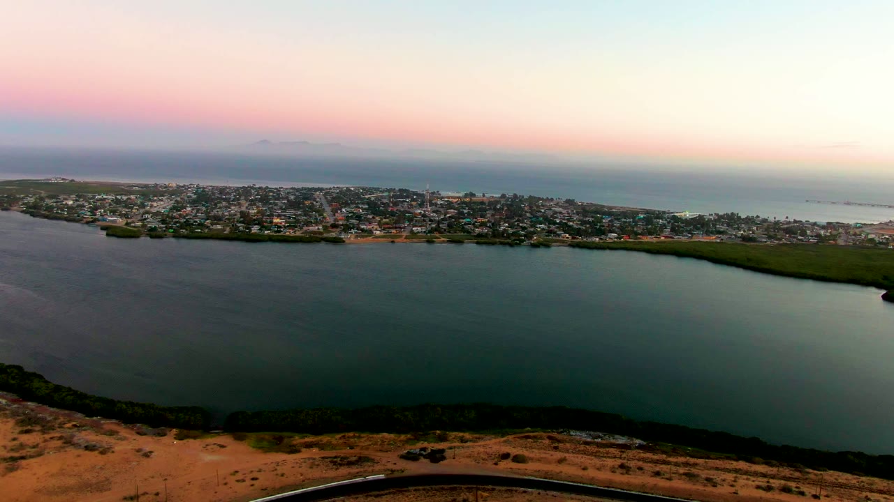 Wide aerial sunset view of bay water with a town and ocean beyond.