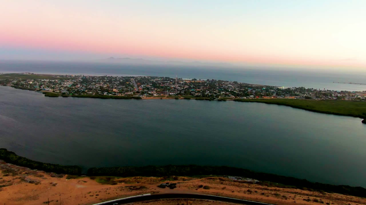 Aerial sunset view of San Carlos showing the bay across the lower frame, the coastal town stretched along the far shore, ocean water behind it, soft pink sky, and no visible people.