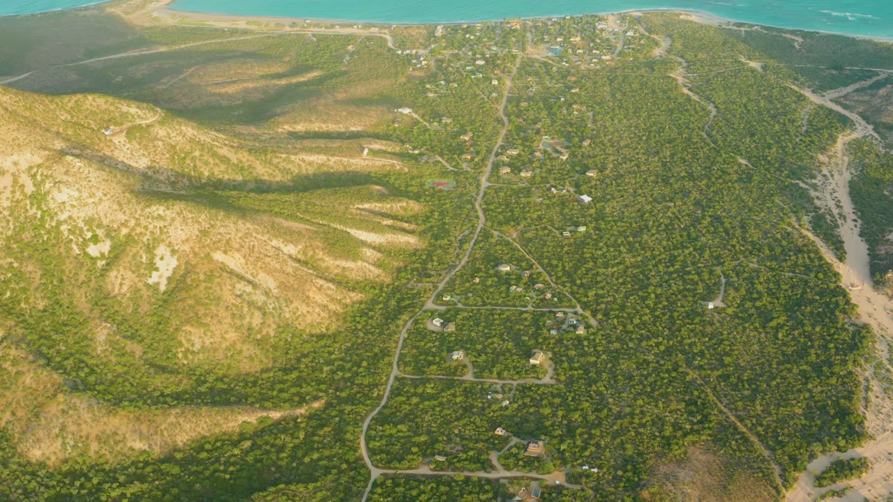 Aerial view over Cabo Pulmo with turquoise water along the top edge, dry hills to the left, green vegetation, winding roads, scattered buildings, and no visible people.