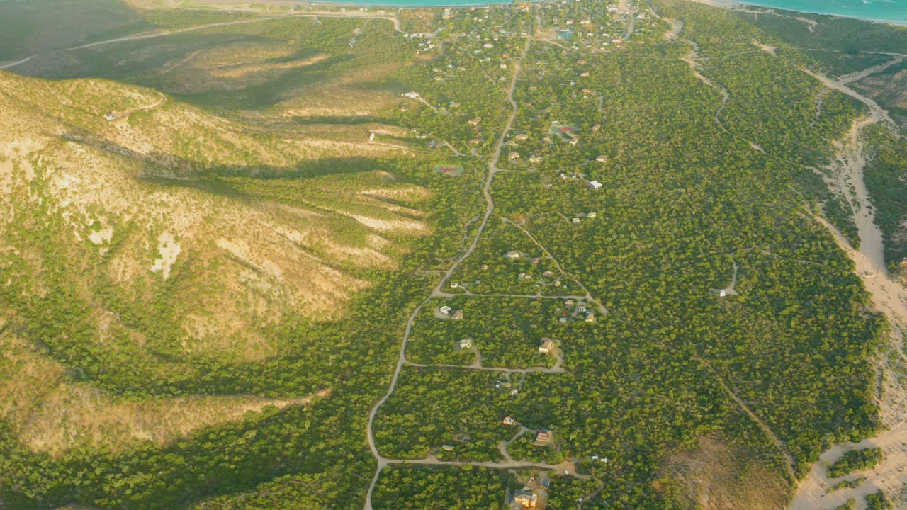 Drone view of green hills, pale slopes, roads, buildings, and a sandy wash.