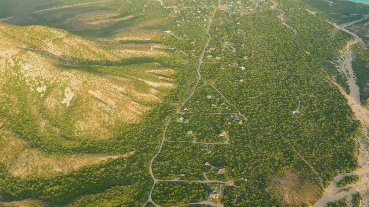 High aerial view over Cabo Pulmo with coastal water still visible near the top, a central road descending through scattered buildings, forested hills, pale dry slopes, and no visible people.