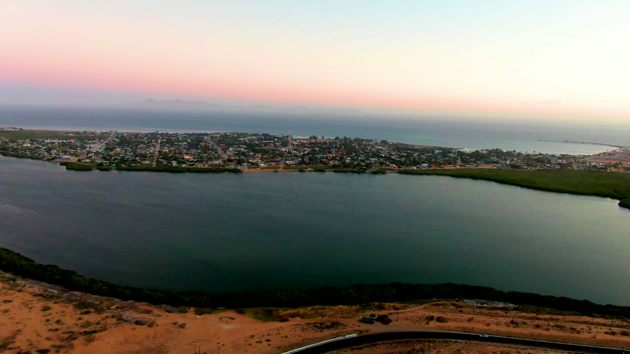 Aerial dusk view of San Carlos, Baja California Sur, with bay water in the foreground, a coastal town across the shore, ocean on the horizon, a curved road below, and no visible people.