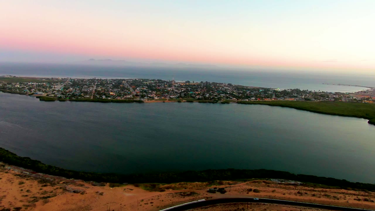Wide aerial view of calm bay water, town buildings, road, and ocean horizon at dusk.