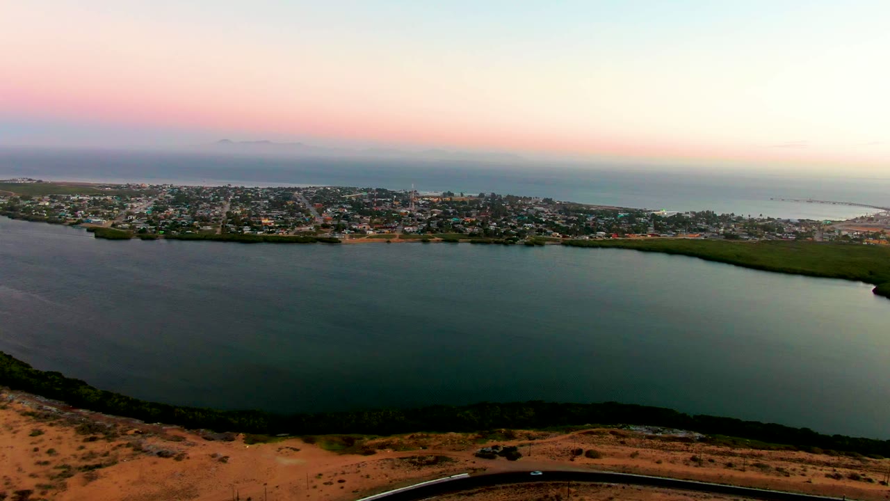 Elevated dusk view in San Carlos, Baja California Sur with broad bay water, a green shoreline strip, town buildings along the far side, distant ocean, and no visible people.