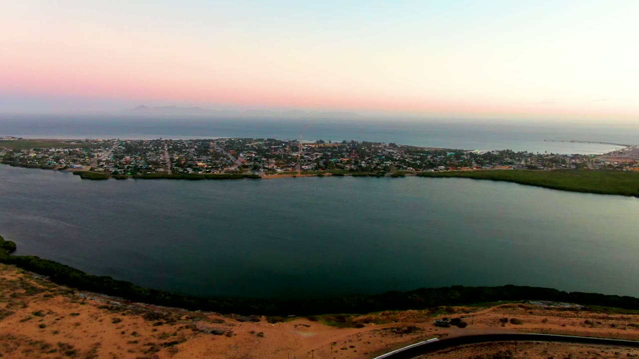 A high view of San Carlos shows the bay in the foreground, the town spread along the shore, darker green land patches near the water, pale sea beyond, and no visible people.