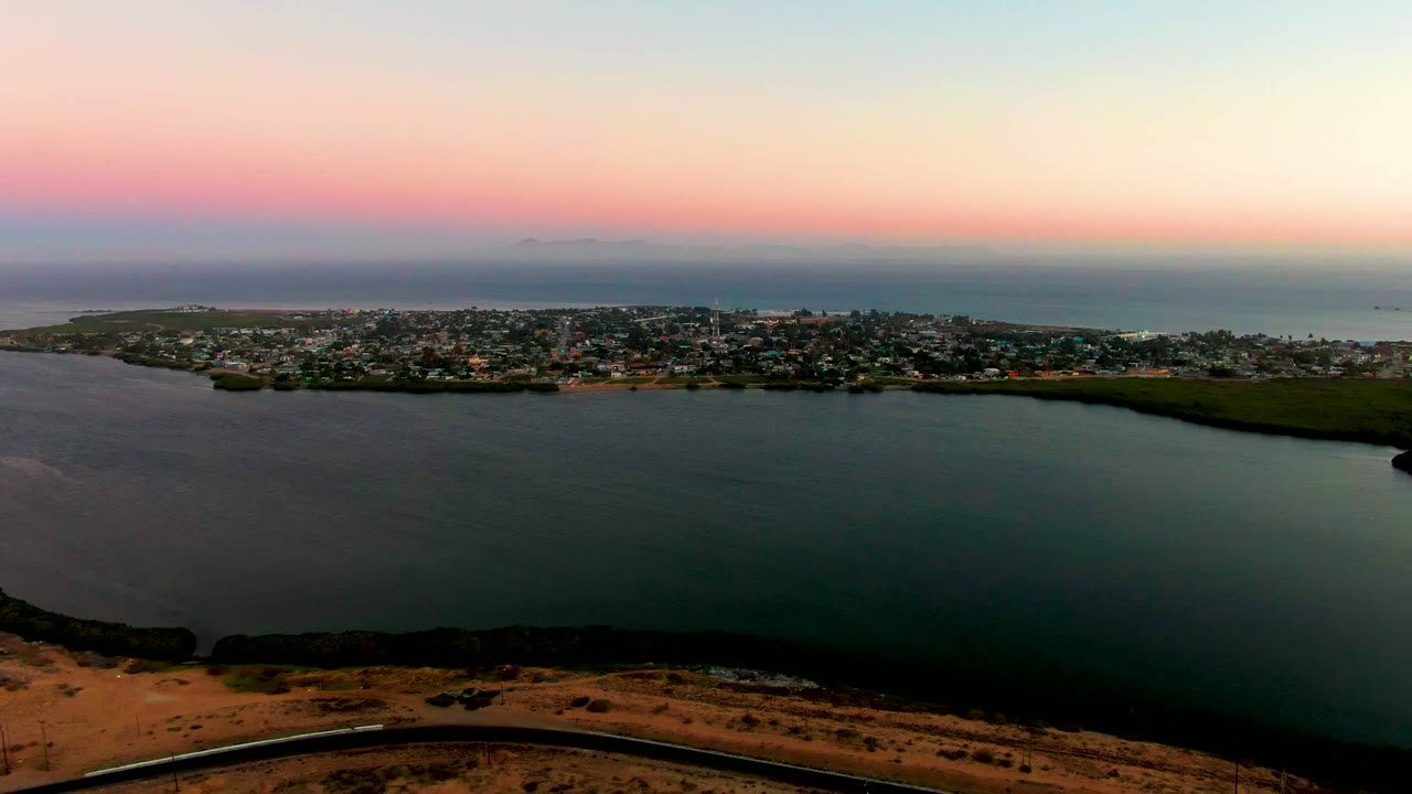 Wide elevated view over San Carlos, Baja California Sur, with dark bay water in front, the coastal town across the shore, a pink dusk sky, and no visible people.