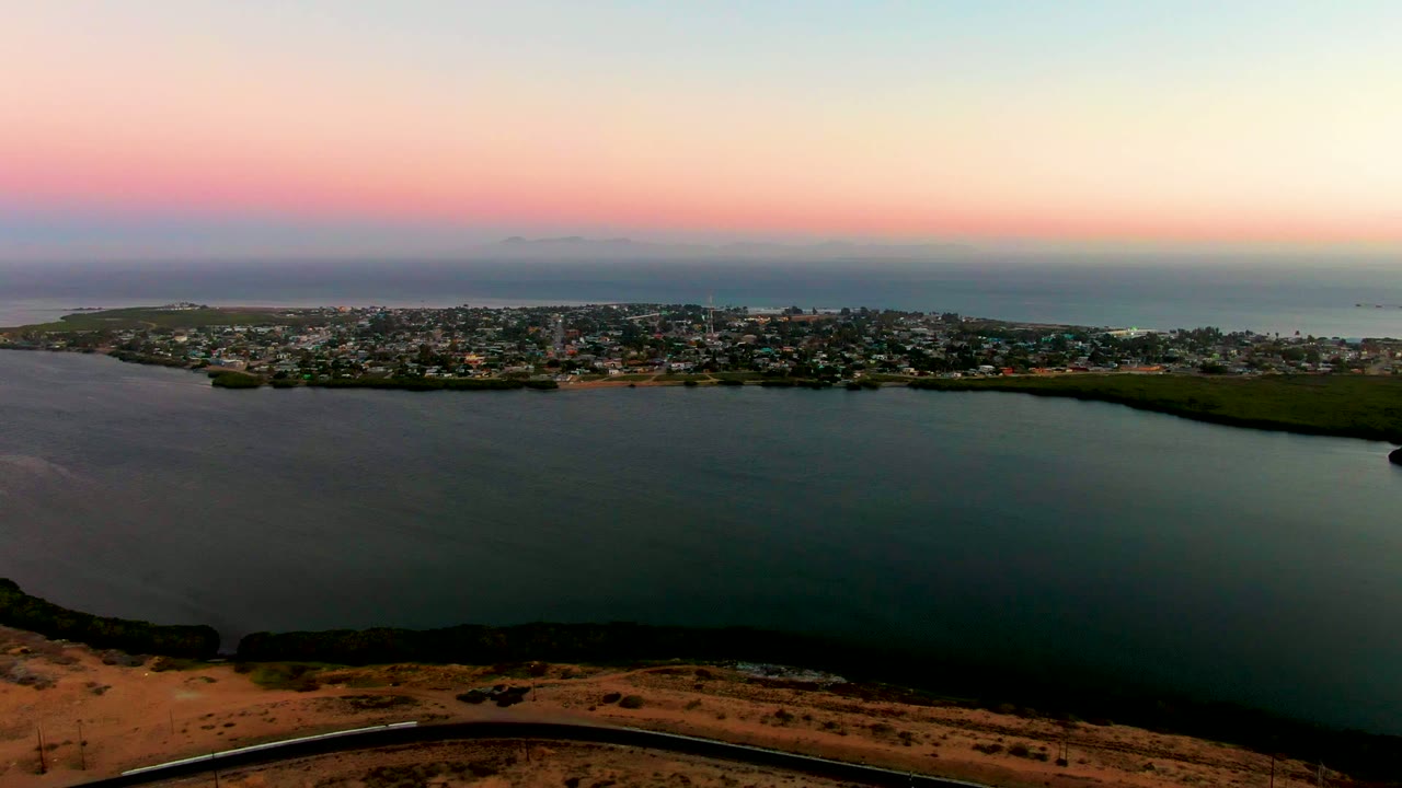 Aerial dusk frame of calm bay water, town shoreline, and foreground road.
