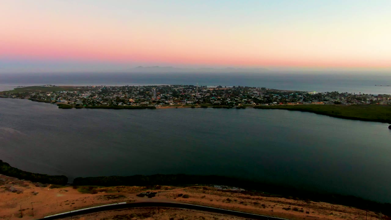 San Carlos, Baja California Sur appears across the bay with clustered buildings, a dark shoreline edge, open ocean beyond, soft pink dusk light, and no visible people.