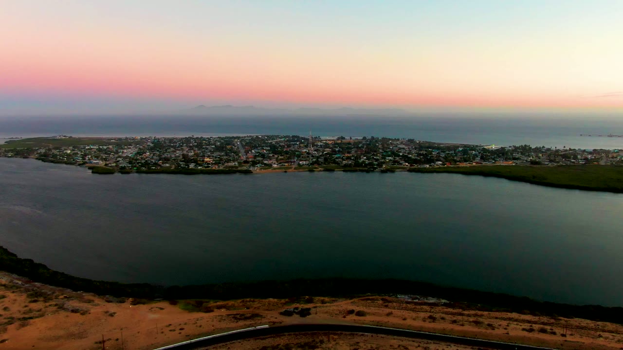 Opening frame of San Carlos, Baja California Sur from an elevated vantage, with dark water in front, the shoreline town across the middle, a pink-blue sunset sky, and no visible people.