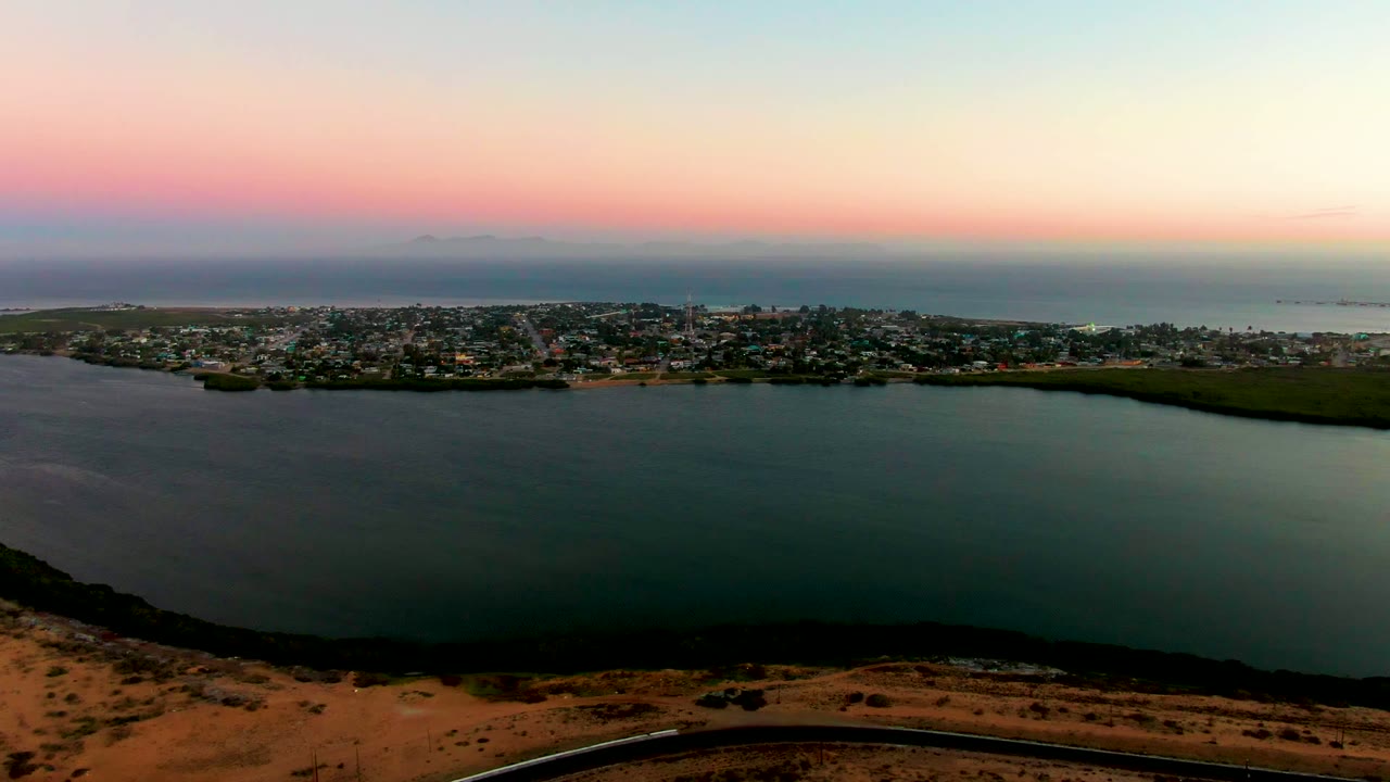 An elevated view of San Carlos shows clustered buildings along the shoreline, broad water below, faint distant ocean beyond, and no visible people.